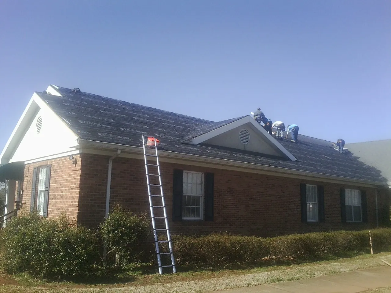 Crew tearing shingles off a brick ranch roof with a ladder set — Emergency Roof Repair work by our crew in Rock Cave