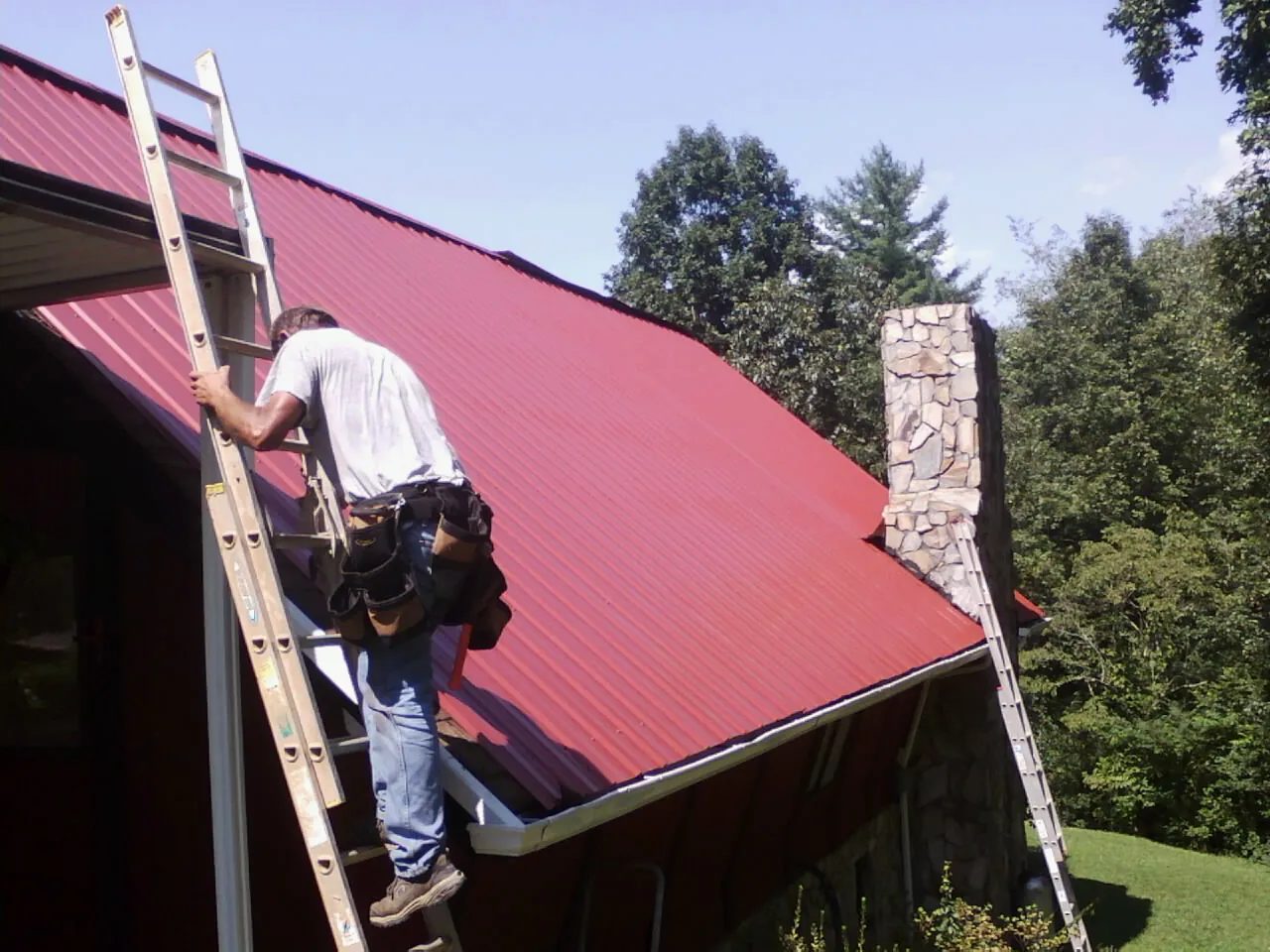 Roofer on a ladder beside a red metal roof and stone chimney — Soffit & Fascia Repair by Roofing Brothers in Rock Cave